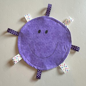 Puede incluir: Un juguete de etiqueta para bebé circular morado con una cara sonriente y cintas adjuntas. El juguete tiene un patrón floral y ojos y boca bordados en negro. Se adjuntan cintas en morado con lunares blancos y blanco con lunares de colores.