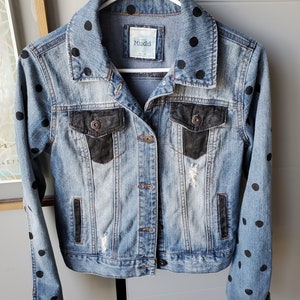 May include: A blue denim jacket with black polka dots on the collar and sleeves. The jacket has black faux leather accents on the pockets and a distressed look. The label on the jacket reads "Mudd".