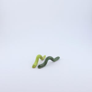 May include: Two small, smooth, worm-shaped figurines. One is a light, bright lime green color, and the other is a darker, forest green shade. Both figures have a curved, undulating body shape and two small, dark dots representing eyes on their heads.