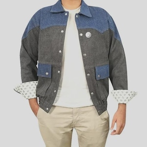 Puede incluir: Chaqueta vaquera bicolor en gris y azul con cierre de botones y dos bolsillos delanteros. La chaqueta tiene un cuello clásico y los puños están doblados, revelando un forro estampado. El modelo lleva una camiseta blanca y pantalones color caqui.