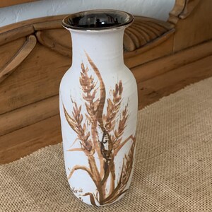 May include: White ceramic vase with a brown rim and a wheat stalk design. The vase has a slightly flared top and a cylindrical body. The wheat stalks are painted in shades of brown and beige, adding a rustic touch to the decor.