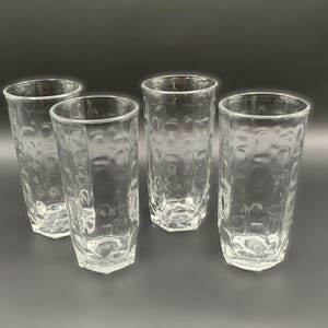 May include: Four clear, textured drinking glasses. Each glass has a tall, cylindrical shape with a hexagonal base. The glass has a dimpled pattern on the exterior. The glasses are arranged on a dark gray surface.