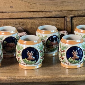 May include: A collection of six ceramic beer steins. Each stein features a circular design with a deer, a house, and foliage. The steins have a cream-colored base with orange and brown accents. The handles are curved.