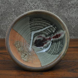 May include: A handmade ceramic bowl with a unique design. The bowl features a segmented pattern with sections of orange, gray-green, and dark gray. White and maroon accents add contrast. The bowl is set on a wooden surface.