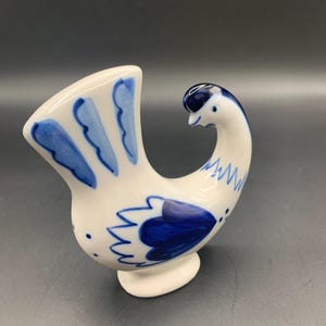 May include: A white ceramic bird figurine with blue floral and feather-like designs. The bird has a curved neck and a small, round base. The tail feathers are fanned out, and the overall design is whimsical and decorative.