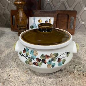 May include: A vintage white ceramic casserole dish with a brown lid and handles. The dish is decorated with a floral pattern in shades of blue, brown, and green. A wooden decorative item is in the background.