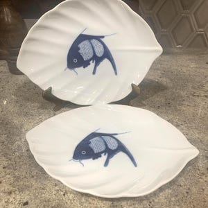 May include: Two white, leaf-shaped ceramic plates with a blue fish design. The fish has a detailed body and a curved tail. The plates are displayed on a countertop, showcasing their decorative and functional design.