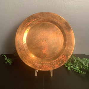 May include: A hammered copper plate with a wide rim and a central design. The plate is displayed on a gold-colored stand. The copper has a warm, reddish-brown hue, and the surface has a textured, dimpled appearance.