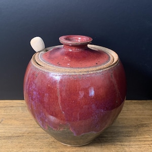 May include: A round, glazed ceramic jar with a lid and a small wooden spoon. The jar is a deep red color with a glossy finish, and the lid has a small knob. The jar sits on a wooden surface.