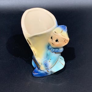 May include: A vintage ceramic figurine of a kneeling child holding a cornucopia-shaped vase. The figurine is painted in shades of blue, yellow, and cream, with gold accents. The child has rosy cheeks and a sweet expression. A decorative piece.
