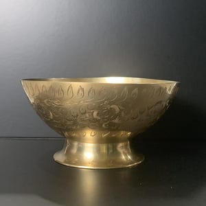 May include: A golden brass bowl with a wide, rounded shape and a pedestal base. The bowl's exterior is adorned with intricate floral and leaf patterns. The polished surface reflects light, highlighting the detailed engravings.