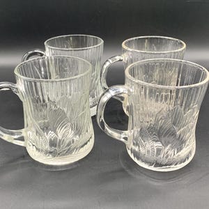 May include: Four clear glass mugs with handles. The mugs have a textured, vertical ribbed design and a floral pattern etched into the glass. The mugs are arranged on a dark surface.
