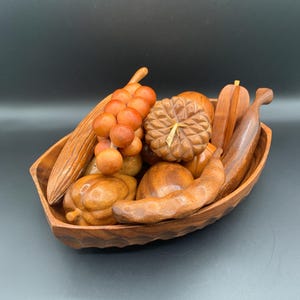 May include: A wooden bowl filled with carved wooden fruit. The bowl contains a bunch of grapes, pumpkins, bananas, and other fruits. The wooden fruit is various shades of brown and orange.