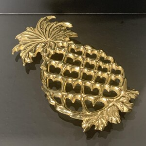 May include: A gold-colored pineapple-shaped decorative object. The pineapple has a textured surface with openwork detailing, and a leafy crown. The object is likely made of metal and is set against a dark background.