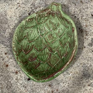 May include: A green ceramic artichoke-shaped dish with a textured surface. The dish has a detailed design with brown accents and a slightly raised rim. The overall shape is oval, and the dish is likely used for serving or display.