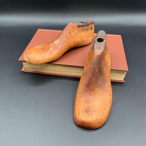 May include: Two antique wooden shoe lasts, with a warm, aged patina. The lasts are resting on a closed, brown book. The shoe forms have a hole at the top and are marked with numbers.