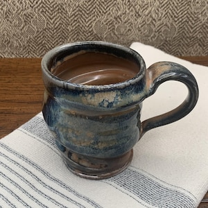 May include: A handmade ceramic mug with a dark blue, brown, and beige glaze. The mug has a rounded body, a sturdy handle, and sits on a small base. The mug is on a white and blue striped cloth.