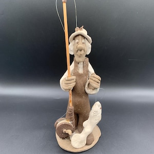 May include: A ceramic figurine of a fisherman holding a fishing rod and reel, with a fish on the line. The figure is wearing a hat and overalls. The color palette is primarily brown and beige.