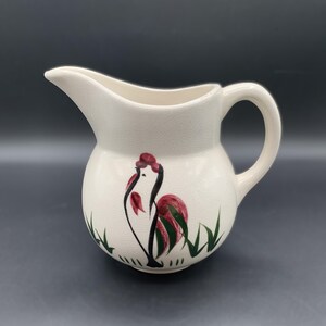 May include: Cream-colored ceramic pitcher with a painted rooster design. The rooster is depicted in red, black, and green, with green grass accents at the base. The pitcher has a curved handle and a spout for pouring.