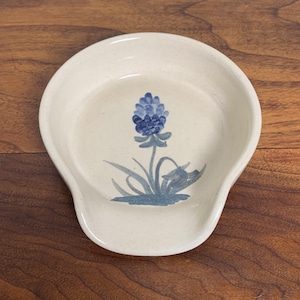 May include: Off-white ceramic spoon rest with a blue floral design. The spoon rest has a rounded shape with a handle rest. The floral design features a blue flower with green leaves and stems.