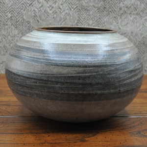May include: A round ceramic vase with horizontal bands of gray, brown, and white. The vase has a wide opening and sits on a wooden surface. The color palette is neutral, making it suitable for various decor styles.