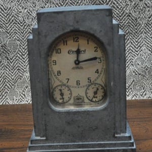 May include: A vintage gray metal clock with a rectangular art deco design. The clock face is cream-colored with black numerals and hands. The word "Fawird" is printed on the clock face.