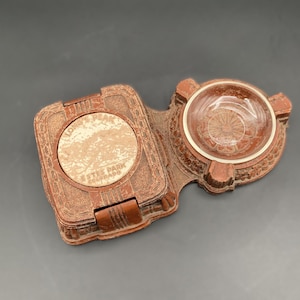 May include: A vintage brown ashtray with a hinged lid and a clear glass bowl. The lid features a circular design with the text "Longs Peak Estes Park Colorado". The ashtray has an ornate, detailed design.