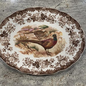 May include: Oval serving platter with a brown floral border and a central image of two pheasants in a natural landscape. The platter has a scalloped edge and is primarily white with brown accents.