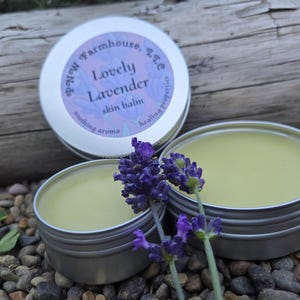 Puede incluir: Tres pequeños botes plateados de bálsamo para la piel con un tono verde claro, acompañados de ramitas de lavanda morada. Un bote está abierto, revelando el bálsamo, y otro tiene una tapa con el texto "Lovely Lavender skin balm". El fondo incluye rocas y madera.