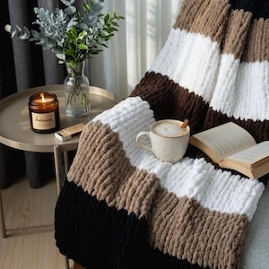 Könnte beinhalten: Eine grob gestrickte Decke mit breiten horizontalen Streifen in Schwarz, Weiß und Hellbraun. Die Decke liegt über einem Stuhl und trägt eine Keramiktasse mit einem Getränk sowie ein aufgeschlagenes Buch mit cremefarbenen Seiten.