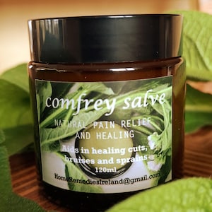 Könnte beinhalten: Ein braunes Glas mit schwarzem Deckel, beschriftet mit "comfrey salve". Das Etikett enthält Informationen über natürliche Schmerzlinderung und Heilung für Schnitte, Prellungen und Verstauchungen. Das Glas enthält 120 ml Produkt, umgeben von grünen Blättern.
