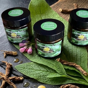Könnte beinhalten: Drei Gläser Comfrey Salbe mit schwarzen Deckeln und grünen Etiketten, die den Text "Double Infused Comfrey" und "Natural Pain Relief and Healing" tragen. Die Gläser sind von grünen Blättern, rosa Blüten und getrockneten Wurzeln umgeben, was auf ein natürliches Heilmittel hindeutet.
