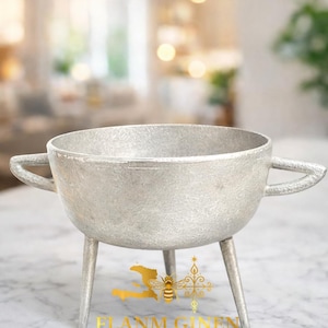 May include: A silver-coloured metal bowl with three legs and two handles. The bowl has a textured surface and is designed to hold candles. The text "FLANM GINEN" is visible on the front, along with other text.