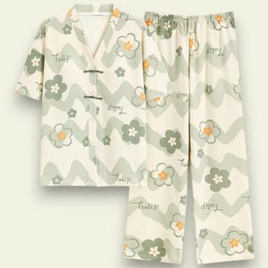 May include: A two-piece pajama set in a cream color with a floral pattern. The set includes a short-sleeved top with a V-neck and frog closures, and matching pants. The design features green and orange flowers and the word "Tulip".