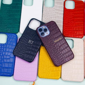 Puede incluir: Una colección de fundas de teléfono coloridas con un patrón texturizado similar a la piel de cocodrilo. Las fundas son de varios colores, incluyendo azul, verde, rojo y morado. Algunas fundas tienen iniciales o nombres personalizados.