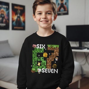 May include: A black sweatshirt featuring a pixelated design with the words "SIX SEVEN" and Minecraft-themed graphics. The design includes blocks, a pig, and other game elements. The sweatshirt is worn by a smiling child.