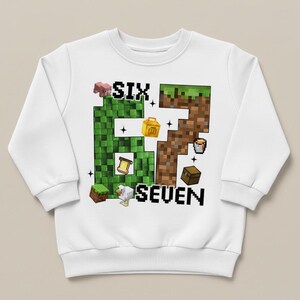 May include: White sweatshirt with a pixelated "6" and "7" design. The "6" is green and the "7" is brown, with various pixelated images of a pig, chicken, chest, and other objects. The word "SIX" is above the "6" and "SEVEN" is below the "7".