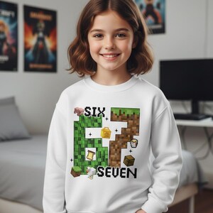 May include: A white sweatshirt with a pixelated "67" design in green and brown, with the words "SIX" and "SEVEN" in black. The design includes pixelated images of a pig, a chest, a chicken, and other game elements. The sweatshirt is worn by a young person.
