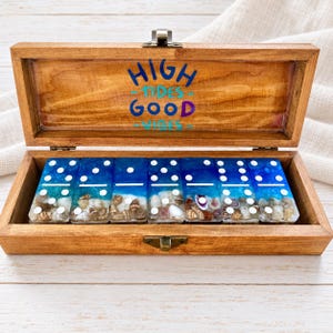 Könnte beinhalten: Eine Holzschatulle mit Metallverschluss enthält sieben Dominosteine aus Kunstharz. Auf der Innenseite des Deckels steht der Text HIGH TIDES GOOD VIBES. Jeder Dominostein hat einen blauen Farbverlauf, weiße Punkte und Muscheln.