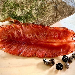 Op de afbeelding: Een decoratieve, blad-vormige hars tray in rood en goud. De tray is versierd met goudvlokken en staat op een lichtgekleurd oppervlak, vergezeld van kleine dennenappels. Een met mos bedekte steen is op de achtergrond.