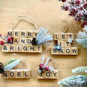 May include: Christmas ornaments made from wooden blocks spelling festive phrases. The ornaments, with twine hangers, feature "Merry and Bright", "Let it Snow", "Noel", and "Joy". They are decorated with faux berries, pinecones, and frosted greenery, set on a light brown surface.