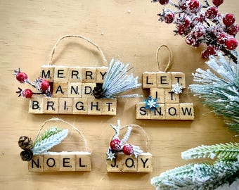 May include: Christmas ornaments made from wooden blocks spelling festive phrases. The ornaments, with twine hangers, feature "Merry and Bright", "Let it Snow", "Noel", and "Joy". They are decorated with faux berries, pinecones, and frosted greenery, set on a light brown surface.