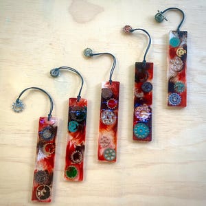 Op de afbeelding: Vijf rechthoekige harsachtige bladwijzers met ingebedde tandwielen en kleurrijke accenten. Elke bladwijzer heeft een unieke opstelling van metalen tandwielen in zilver, brons en koper, tegen een rode en witte achtergrond. Een klein tandwiel is bevestigd aan een zwart koord.