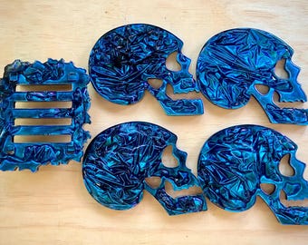 Peut inclure: Ensemble d'objets décoratifs en forme de crânes bleus et un élément rectangulaire similaire. Les crânes présentent une finition texturée et brillante, avec des nuances de bleu et de turquoise. L'ensemble est disposé sur une surface en bois clair.