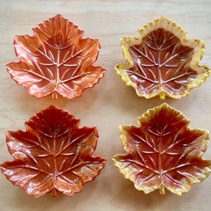 May include: Four leaf-shaped dishes in shades of orange and gold. Two dishes are solid orange, while the other two have gold rims. The dishes are arranged on a light-coloured wooden surface.