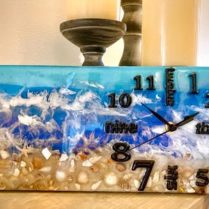May include: A rectangular clock with a beach-themed design. The clock face features a blue and white resin wave pattern with embedded seashells and sand. Black numerals and hands complete the design.