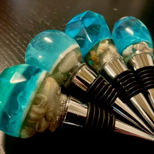 May include: A selection of decorative wine bottle stoppers. Each stopper has a clear, blue resin top, incorporating seashells. The stoppers have a silver metal base and a black rubber seal. The design is reminiscent of the ocean.
