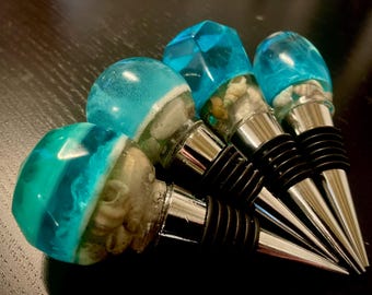 May include: A selection of decorative wine bottle stoppers. Each stopper has a clear, blue resin top, incorporating seashells. The stoppers have a silver metal base and a black rubber seal. The design is reminiscent of the ocean.