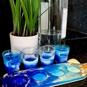 May include: A set of four shot glasses with blue and white swirling designs, displayed on a matching serving tray. A bottle of liquor and a potted plant are in the background. Seashells are placed near the tray.