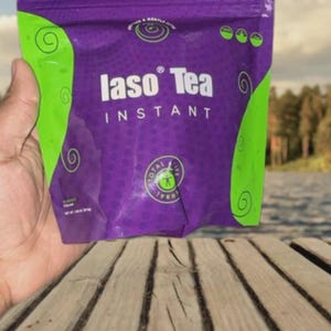 May include: A purple and green packet of "Iaso Tea Instant" held in a hand. The packet has green swirls and the words "Iaso Tea" in white. The background shows a lake and trees.